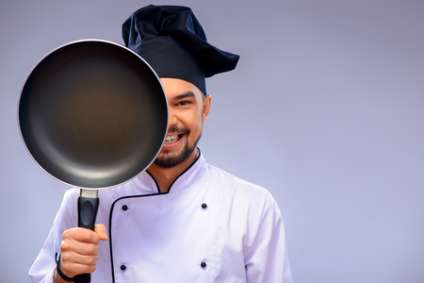 He is a best chef ever. Closeup portrait of handsome cook covering a half of his face with a frying pan and smiling wh