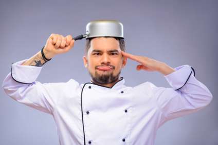 Funny cook. Closeup portrait of handsome cook making faces with a small pan on his head while standing over grey background with copy space