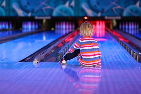 Adorable little child, blonde toddler girl, sitting on the floor at bowling club waiting for a strike - weekend family leisure concept